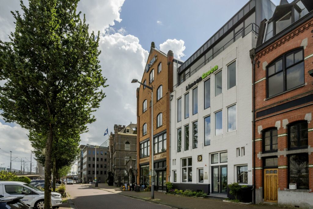 Gevels van verschillende kantoorpanden aan de De Ruijterkade in Amsterdam, met bomen en geparkeerde auto's op de voorgrond.